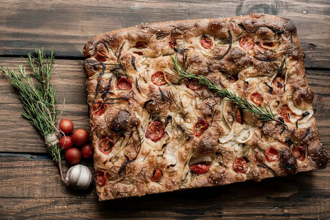 Fresh rosemary focaccia on a rustic wooden table with garlic and cherry tomatoes